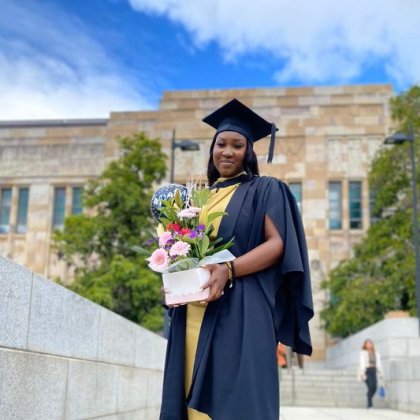 A young African woman stands in graduation cap and gown and holding flowers outside UQ's sandstone Great Court.